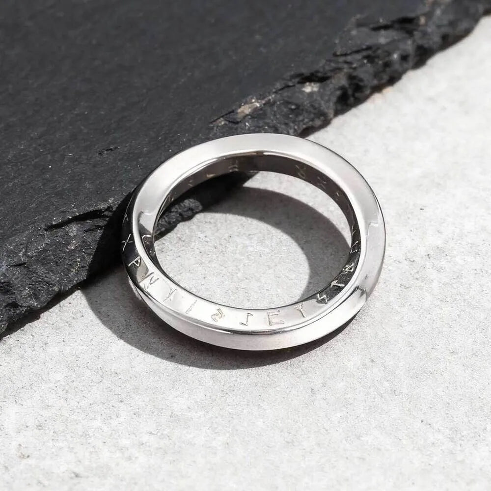 Ein schmaler, silberfarbener Möbius-Schleifen-Ring aus Edelstahl, flach liegend auf einer dunklen Schieferplatte präsentiert.