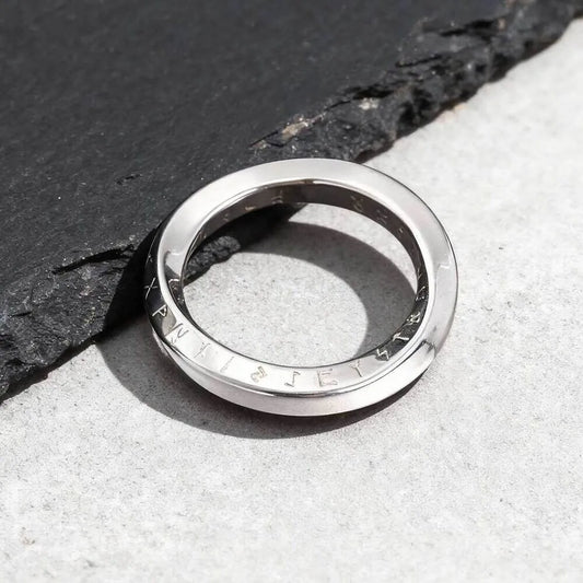 Ein schmaler, silberfarbener Möbius-Schleifen-Ring aus Edelstahl, flach liegend auf einer dunklen Schieferplatte präsentiert.