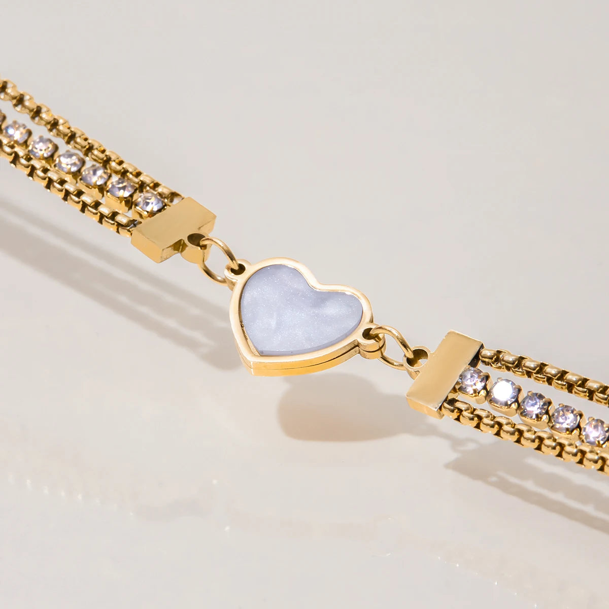 Nahaufnahme des filigranen Schmuckstücks: Mehrreihiges goldenes Strassarmband mit einem schimmernden Herz-Inlay, diagonal fotografiert.