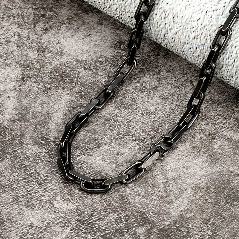 Nahaufnahme der schwarzen, matten Edelstahl-Gliederkette. Der Fokus liegt auf der Vintage-Textur der dicken, rechteckigen Kettenglieder, liegend auf einem dunklen, steinernen Untergrund.