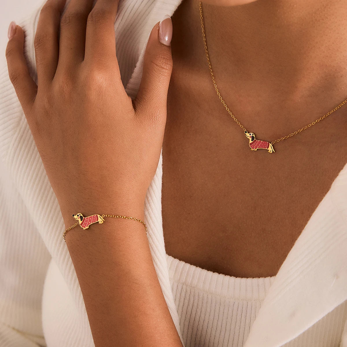 Eine Frau trägt ein passendes Schmuckset bestehend aus einer Halskette und einem Armband aus 18K goldvergoldetem Edelstahl, beide mit einem kleinen Dackel-Anhänger, der mit rosa Emaille-Details verziert ist.