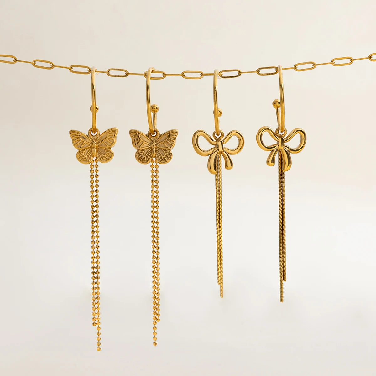 Produktübersicht der Kollektion: Vier unterschiedliche, lange Ohrhänger aus vergoldetem Edelstahl mit Schmetterlings- und Schleifen-Motiven, aufgereiht an einer goldenen Gliederkette.