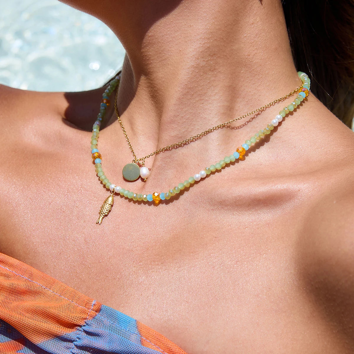 Modell trägt ein sommerliches Halsketten-Set aus einer feinen 18K vergoldeten Edelstahlkette mit rundem Plättchen und einer echten Süßwasserperle, kombiniert mit einer bunten Perlenkette aus hellgrünen, blauen und orangenen Perlen sowie einem goldenen Fisch-Anhänger.