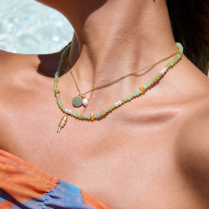 Modell trägt ein sommerliches Halsketten-Set aus einer feinen 18K vergoldeten Edelstahlkette mit rundem Plättchen und einer echten Süßwasserperle, kombiniert mit einer bunten Perlenkette aus hellgrünen, blauen und orangenen Perlen sowie einem goldenen Fisch-Anhänger.