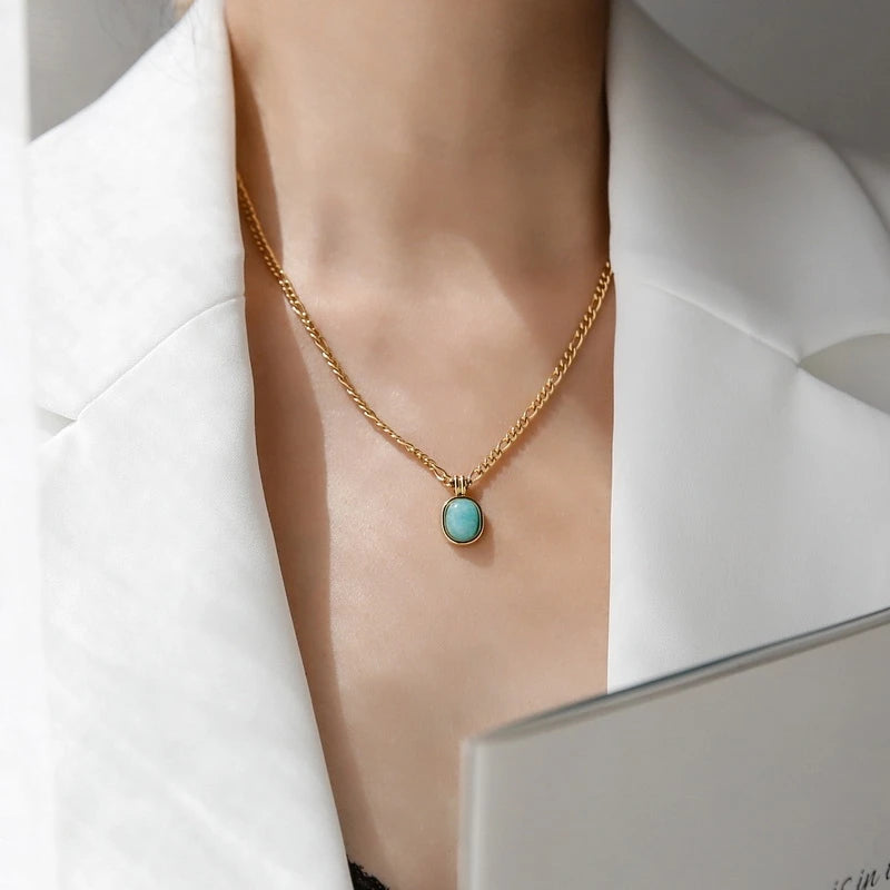 „Nahaufnahme einer Frau, die eine mittellange goldene Kette mit einem ovalen, türkisblauen Anhänger über einem schwarzen Top trägt.“