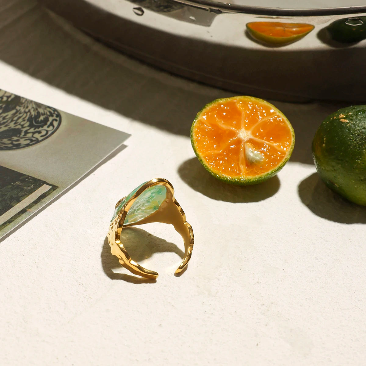 Rückseite eines ovalen Rings aus 18K vergoldetem Edelstahl mit Türkis-Stein und Seestern-Symbol sowie einem Muschel-Symbol, neben einer aufgeschnittenen Orange.