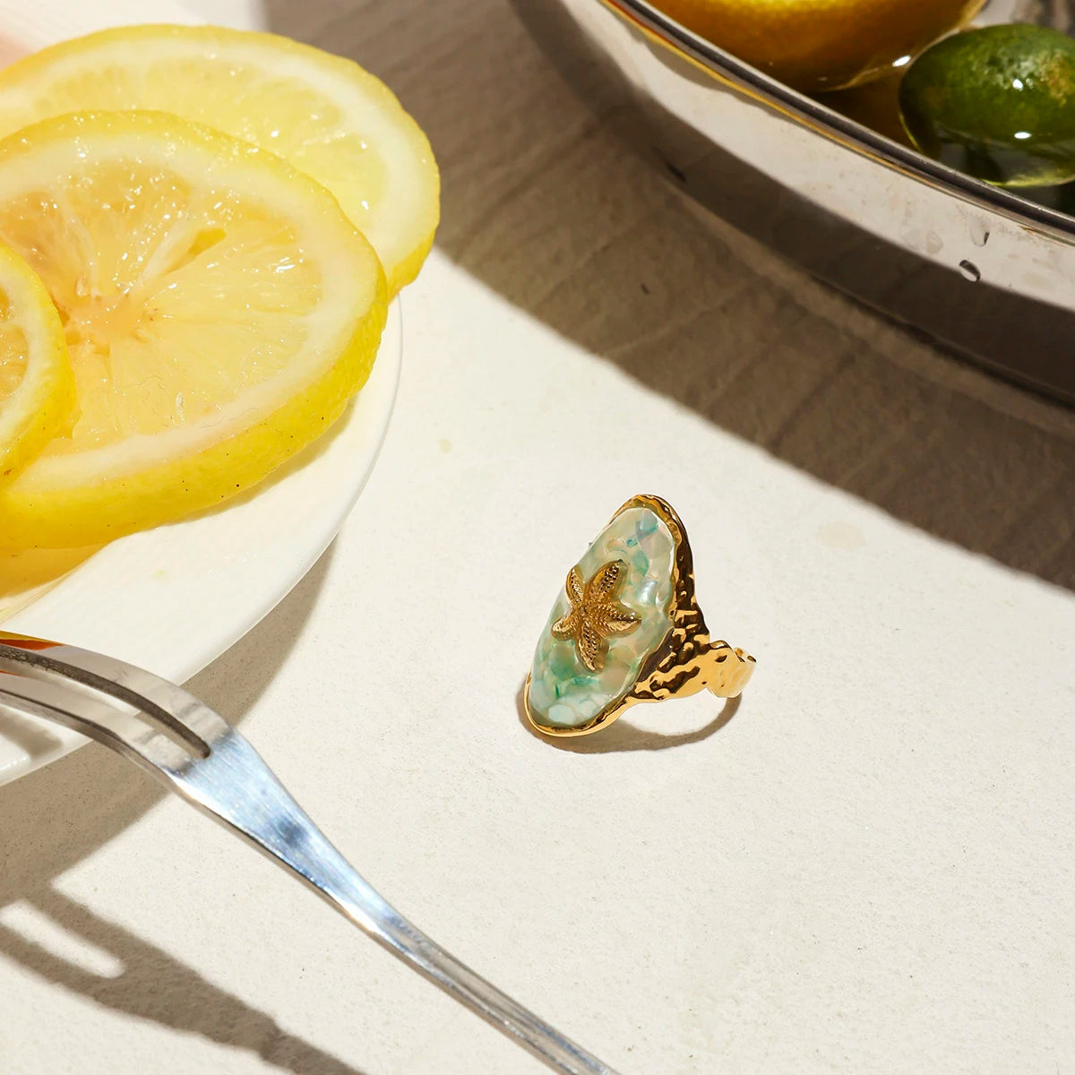 Ein Ring aus 18K vergoldetem Edelstahl mit einem Muschel-Symbol auf einem Tisch, im Hintergrund eine Schale mit Oliven.