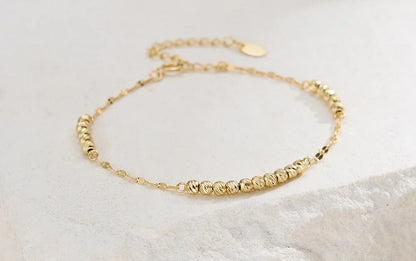 Filigranes, goldfarbenes Armband aus 14K vergoldetem 925er Sterling Silber, verziert mit mehreren kleinen, strukturierten Kügelchen und einer verstellbaren Verlängerungskette, liegend auf einem hellen Stein.