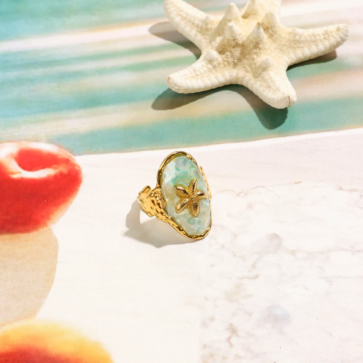 Ovale Ringe aus 18K vergoldetem Edelstahl, einer mit türkisfarbenem Stein und Seestern, der andere mit weißem Muschel-Symbol, auf Eiswürfeln am Strand.