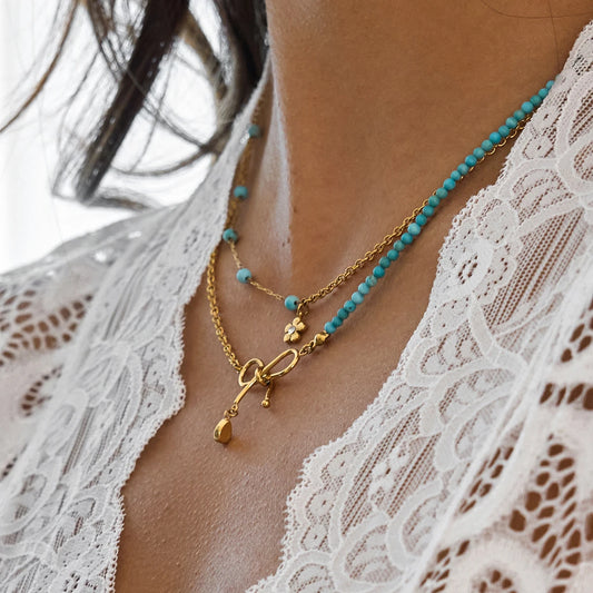 Nahaufnahme einer goldenen Kette aus 18K vergoldetem Edelstahl mit feinen, türkisfarbenen Perlen. Ein verspielter Schleifen-Anhänger mit einem glitzernden Zirkonia-Stein hängt daran