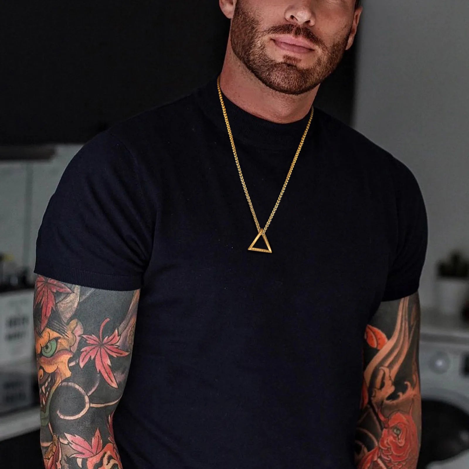 Goldfarbene Edelstahlkette mit kleinem, offenem dreieckigen Anhänger, getragen von einem Mann mit Tätowierung über einem schwarzen T-Shirt.