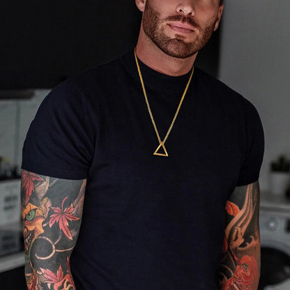 Goldfarbene Edelstahlkette mit kleinem, offenem dreieckigen Anhänger, getragen von einem Mann mit Tätowierung über einem schwarzen T-Shirt.