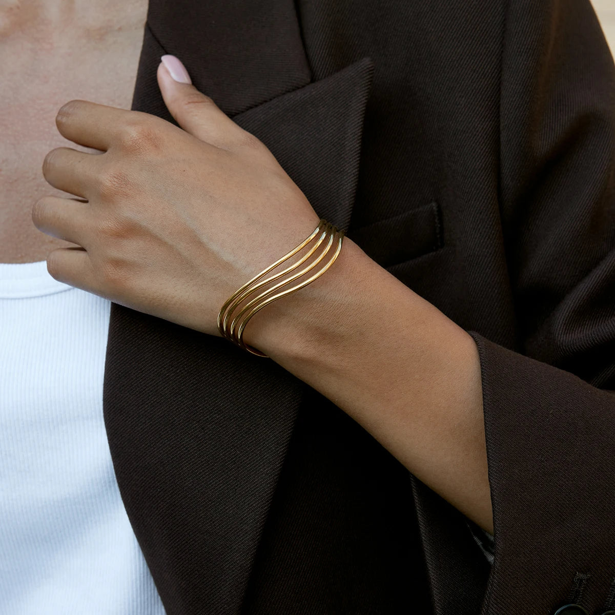 Nahaufnahme am Handgelenk: Eleganter, mehrreihig geschwungener Armreif aus 18K vergoldetem Edelstahl zu einem dunkelbraunen Blazer.