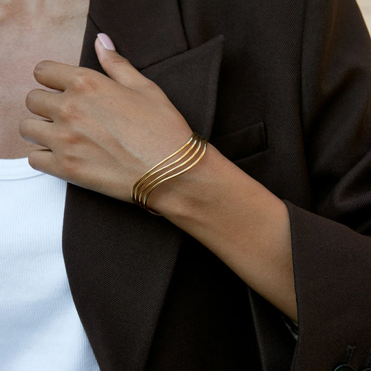 Nahaufnahme am Handgelenk: Eleganter, mehrreihig geschwungener Armreif aus 18K vergoldetem Edelstahl zu einem dunkelbraunen Blazer.