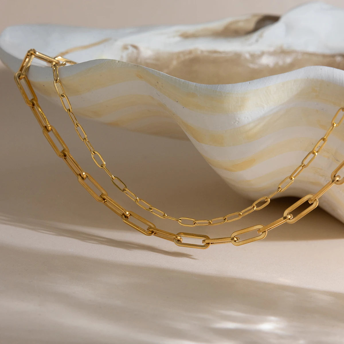 Kunstvolle, sonnenlichtdurchflutete Nahaufnahme des geschichteten goldvergoldeten Edelstahl-Paperclip-Kettensets, platziert in und neben einer hellen, organisch geformten Muschelschale auf einem hellen Hintergrund.