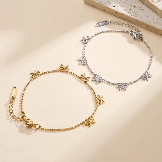 Zwei filigrane Edelstahl-Armbänder mit kleinen Schleifen-Anhängern in Gold (18K vergoldet) und Silber nebeneinander präsentiert.