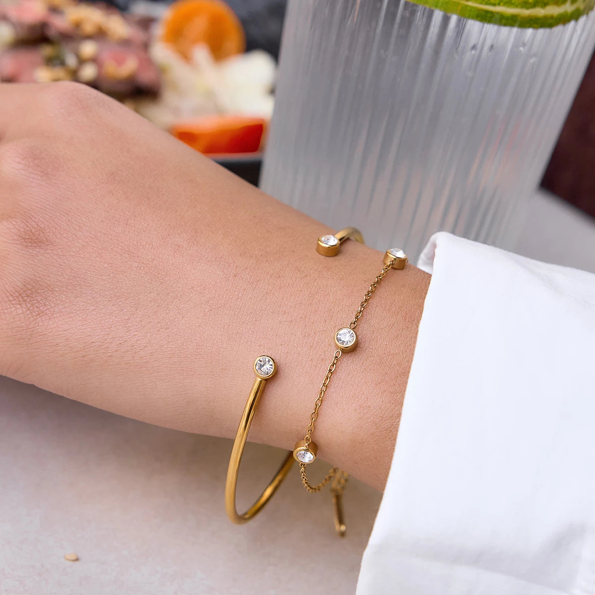 Nahaufnahme eines 2-teiligen Armband-Sets am Handgelenk: Ein offener, 18K vergoldeter Edelstahl-Armreif mit Zirkoniern an den Enden, kombiniert mit einer feinen Goldkette mit runden Kristalleinfassungen.