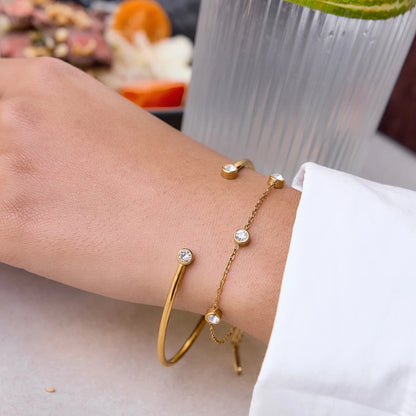 Nahaufnahme eines 2-teiligen Armband-Sets am Handgelenk: Ein offener, 18K vergoldeter Edelstahl-Armreif mit Zirkoniern an den Enden, kombiniert mit einer feinen Goldkette mit runden Kristalleinfassungen.