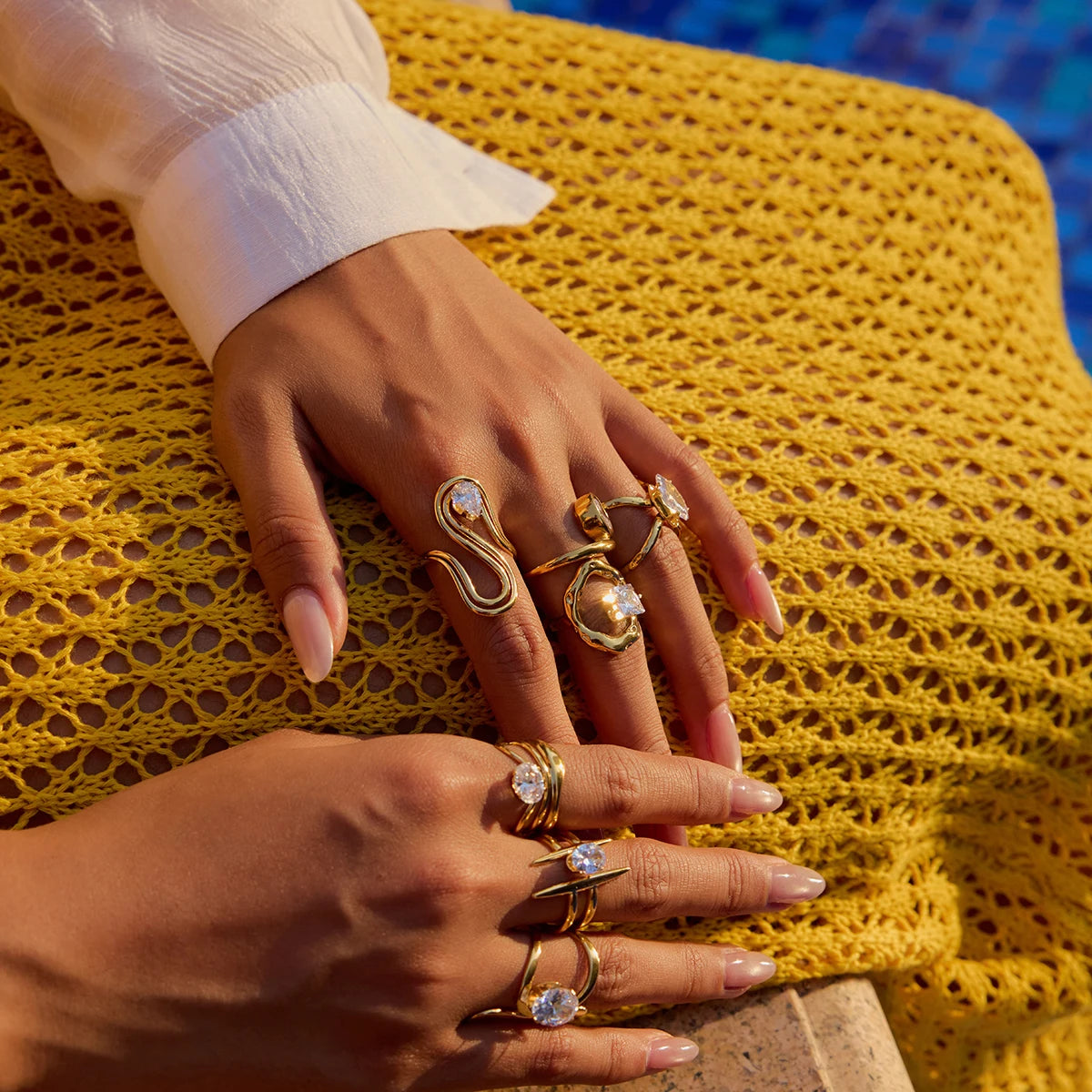 Die Hände einer Person sind mit mehreren goldenen Ringen mit 18K PVD-Vergoldung geschmückt, die über verschiedene Finger verteilt sind. Die Ringe haben ein abstraktes Design und sind mit großen, klaren Zirkonia Steinen besetzt.