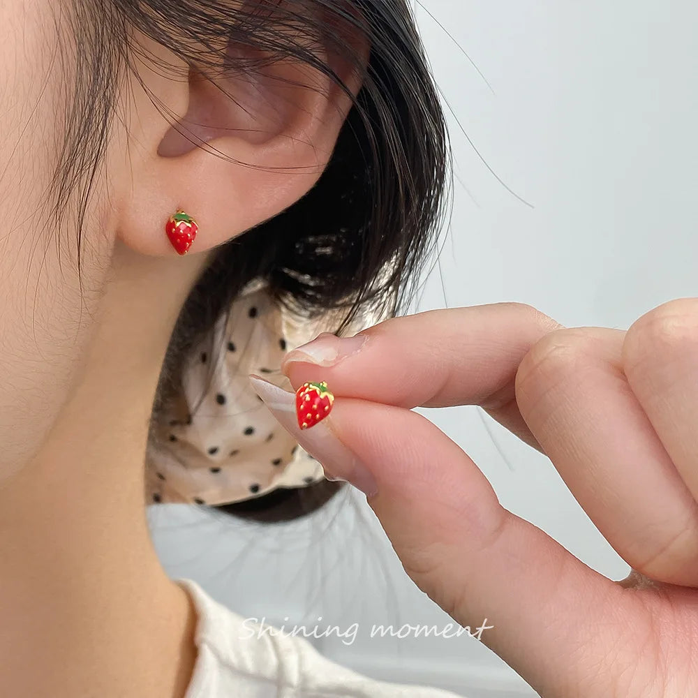 Model trägt den kleinen Erdbeer-Ohrstecker am Ohrläppchen und hält einen weiteren zwischen den Fingern.