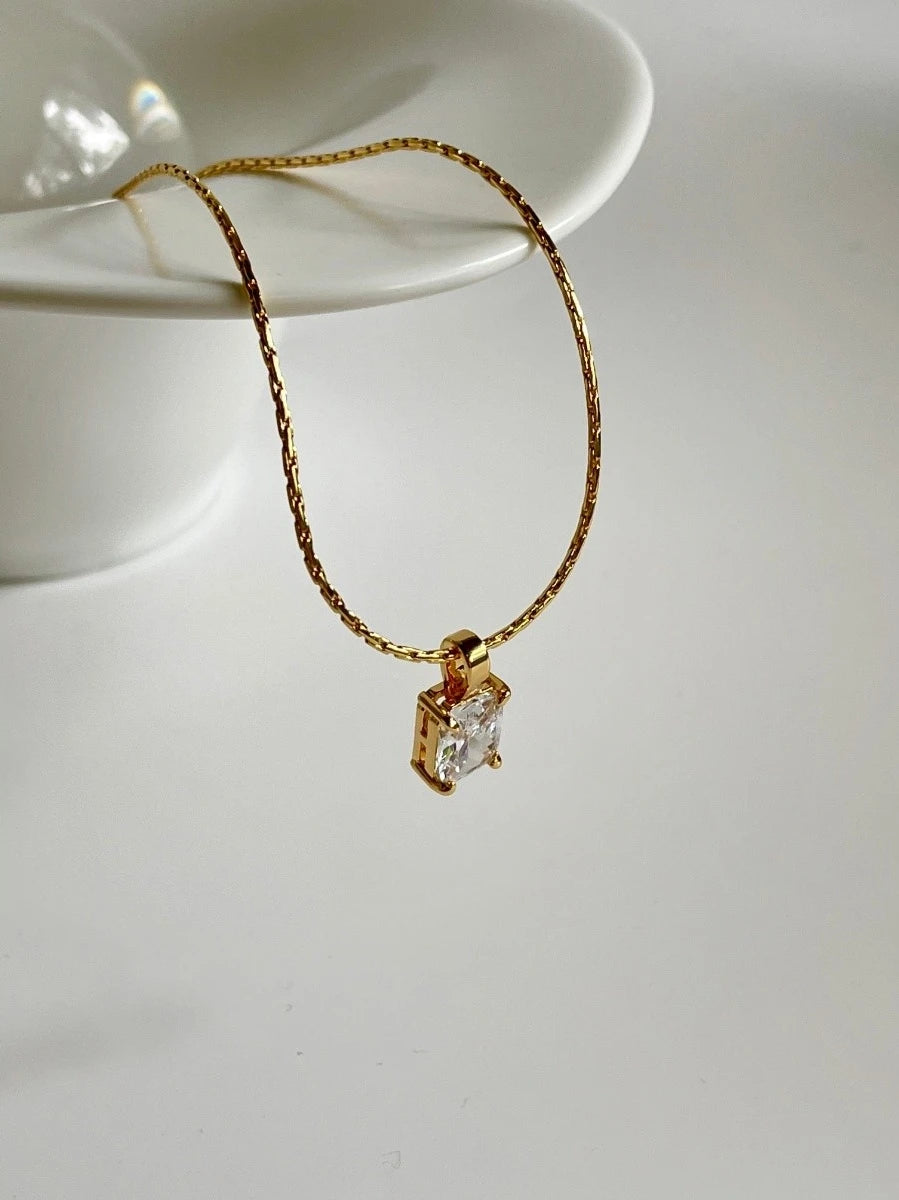 Goldene Kette mit rechteckigem Solitär-Zirkonia-Anhänger, elegant drapiert an einer weißen, wellenförmigen Keramikschale.