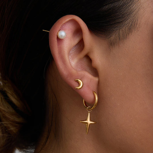 Nahaufnahme vom Ohr einer Frau, das mehrere goldene Edelstahl-Ohrringe mit 18K PVD-Vergoldung trägt. Ein großes Set aus einem Kreolen-Ohrring und einem baumelnden Stern ist im ersten Loch zu sehen. Ein kleiner Halbmond ist im zweiten und eine schlichte, kleine Kugel im obersten Loch platziert.