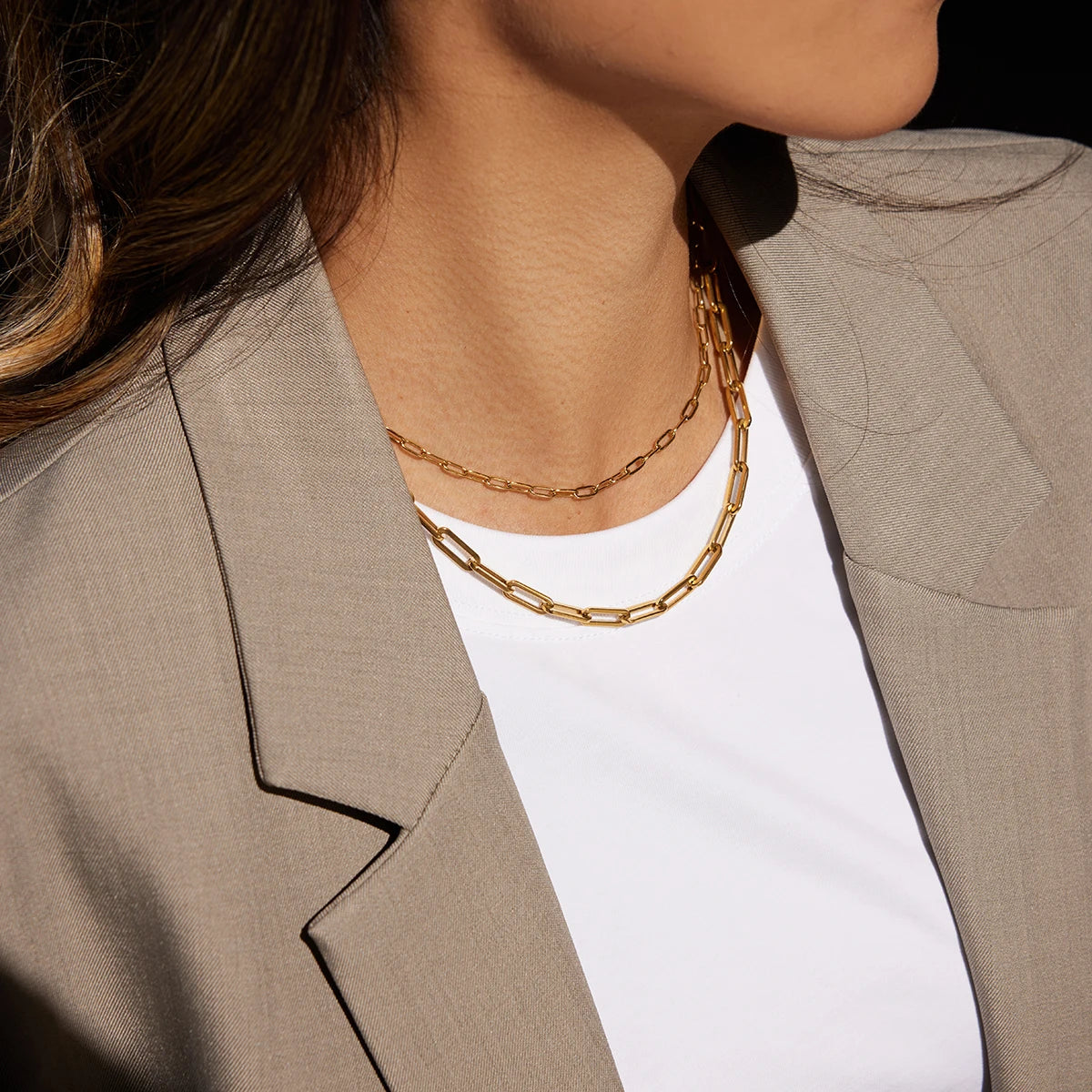 Detailansicht eines geschichteten Halskettensets aus goldvergoldetem Edelstahl am Hals einer Trägerin, das das modern-elegante Paperclip-Design über einem schlichten weißen T-Shirt und einem beige Blazer hervorhebt.