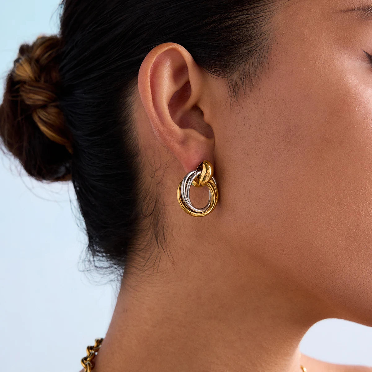 Eine Seitenansicht des Gesichts einer Person, die einen Ohrring aus zwei ineinandergreifenden Ringen trägt. Der obere Ring ist golden, der untere ist silberfarben. Im Hintergrund ist ein Ozean zu sehen.