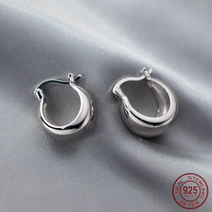 Ein Paar glänzender, kleiner, bauchiger Creolen-Ohrringe (Huggie-Ohrringe) aus 925er Sterlingsilber, liegend auf einem hellgrauen, seidenartigen Stoff. Das 925 Sterling Silver Echtheitssiegel ist in der unteren rechten Ecke sichtbar.