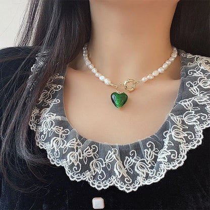 Elegante Perlenkette mit vergoldeter Knebel-Schließe und grünem Herzanhänger zu einem schwarzen Kleid mit Spitzenkragen.