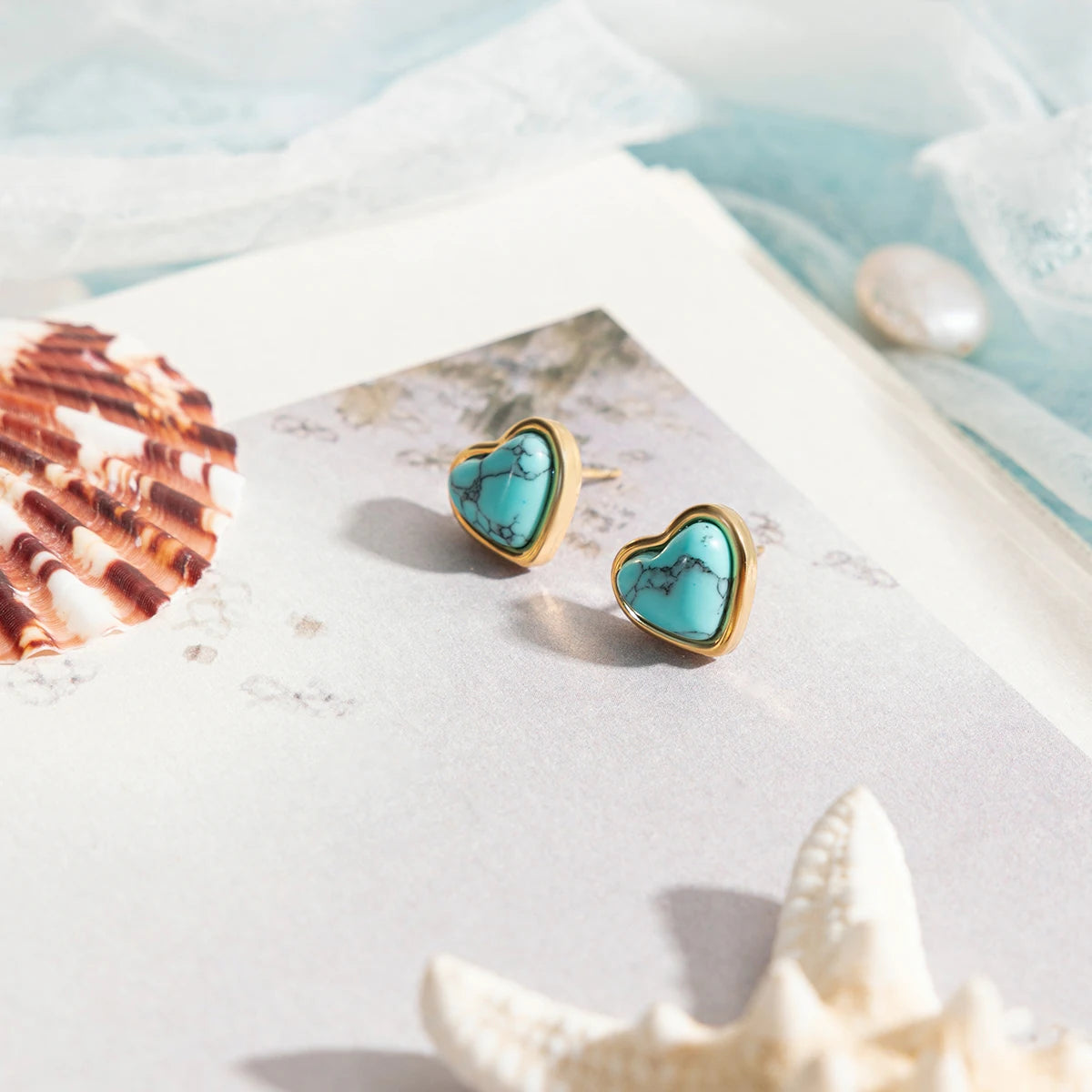 Zwei Paare Herz-Ohrstecker aus 18K vergoldetem Edelstahl mit blauem Türkisstein, auf einem Untergrund mit Muscheln.