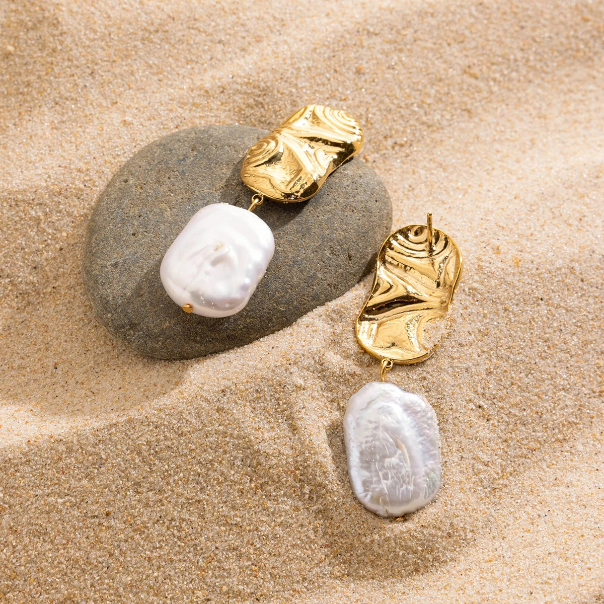 Goldene Ohrringe mit unregelmäßiger Süßwasserperle, drapiert auf Sand und Muscheln. Ein sommerlicher und maritimer Schmuck-Look.