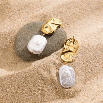 Goldene Ohrringe mit unregelmäßiger Süßwasserperle, drapiert auf Sand und Muscheln. Ein sommerlicher und maritimer Schmuck-Look.