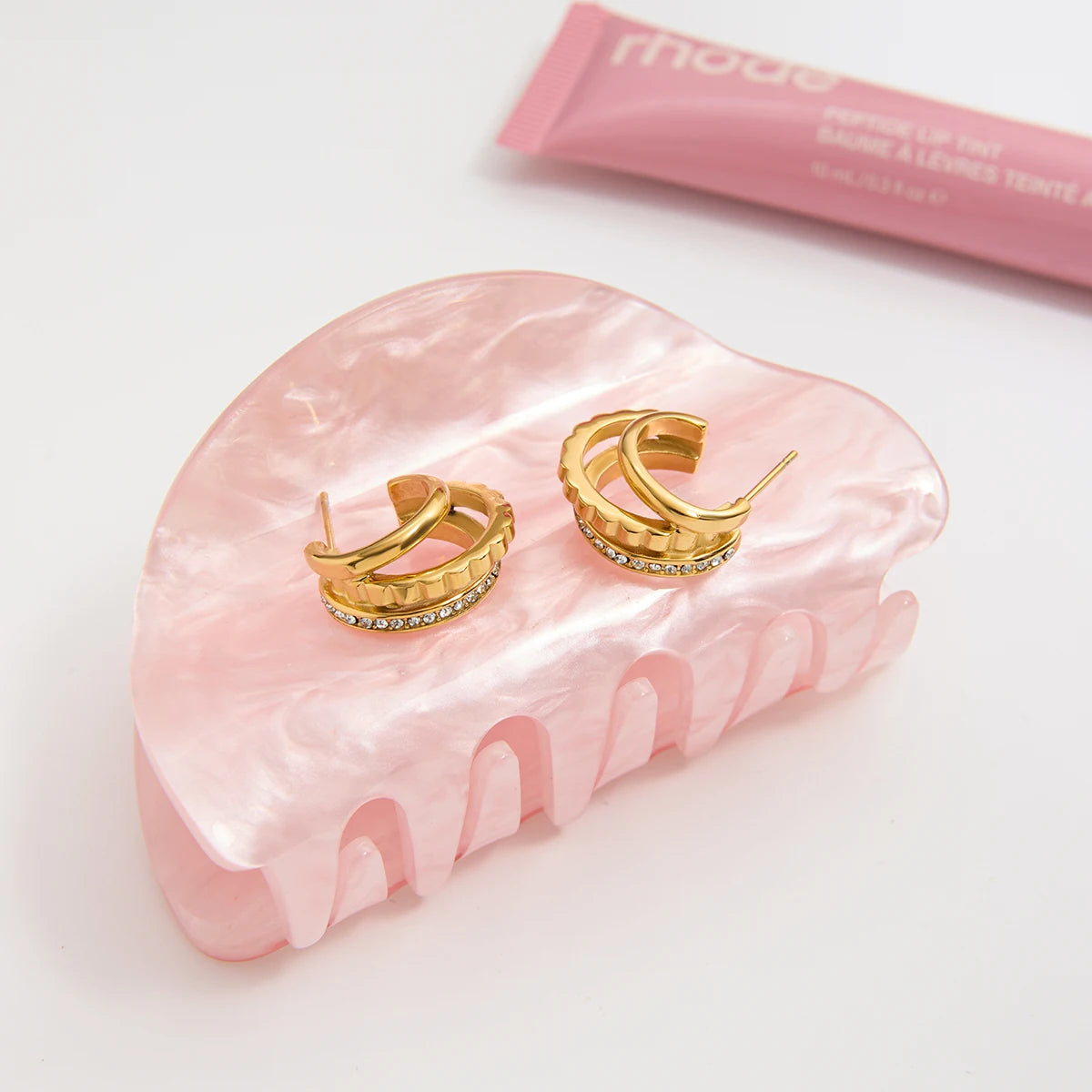 „Ästhetische Produktpräsentation: Die goldenen Creolen liegen dekorativ auf einer rosa marmorierten Haarklammer neben einer Tube Lipgloss.“