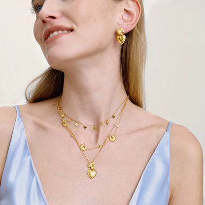 Eine Frau trägt ein mehrlagiges Halskettenset und passende Ohrringe aus 18K goldvergoldetem Edelstahl mit Herz- und Blumendetails.