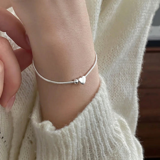 Dezentes Tragebild am Handgelenk einer Frau: Zartes Silberarmband mit kleinem Herz-Charm zu einem hellem Strickpullover