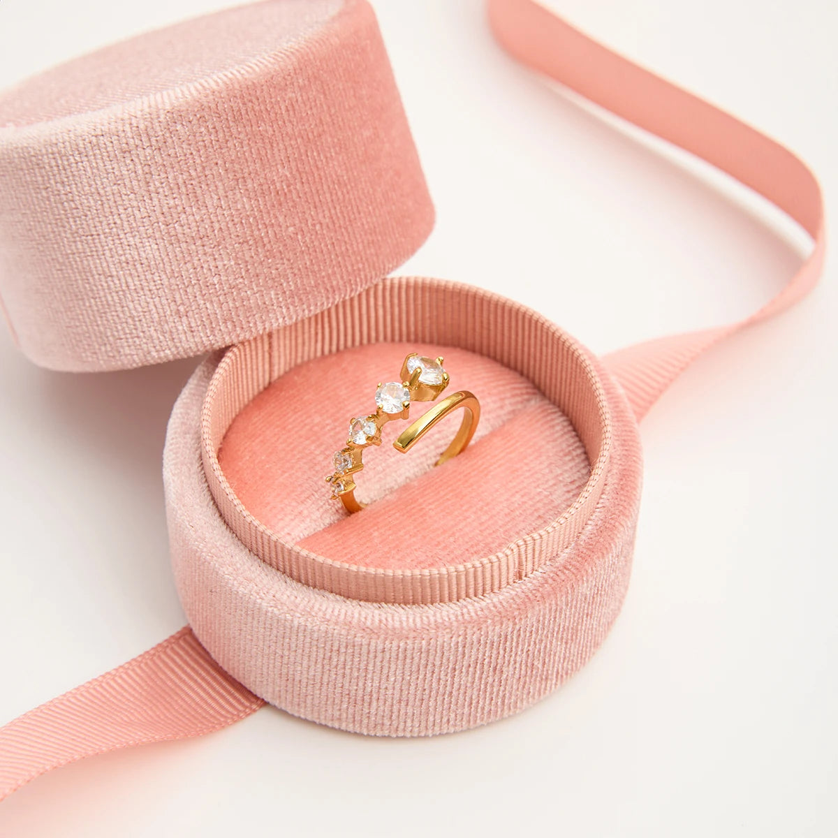 „Der offen gestaltete Goldring mit funkelnden Steinen, elegant präsentiert in einer runden, hellrosa Samt-Schmuckschatulle mit passendem Seidenband.“