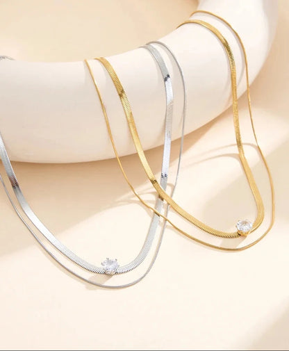 Zweireihige Schlangenketten aus Edelstahl mit Zirkonia-Anhänger, ein Modell in Gold und eines in Silber, drapiert auf einem hellen Podest.