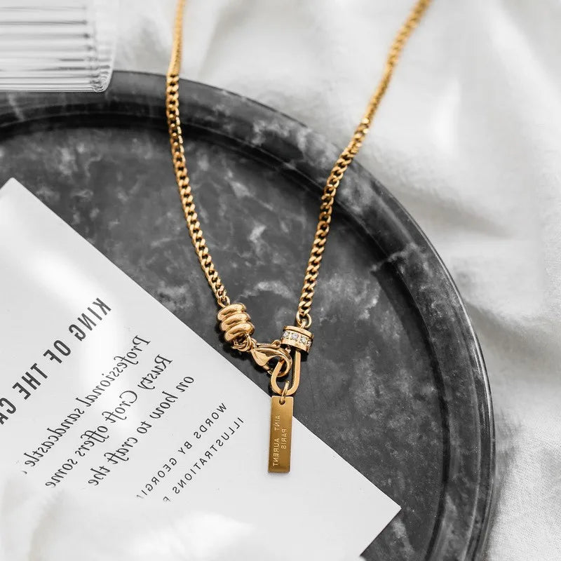 Detailansicht der goldenen Titan-Kette mit Y-Knotenelement und Stäbchen-Anhänger, präsentiert auf einer marmorierten Deko-Schale.