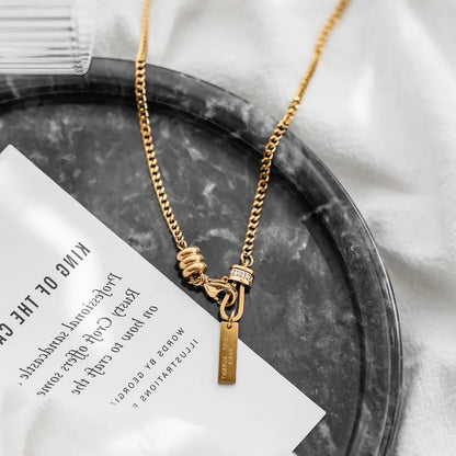 Detailansicht der goldenen Titan-Kette mit Y-Knotenelement und Stäbchen-Anhänger, präsentiert auf einer marmorierten Deko-Schale.