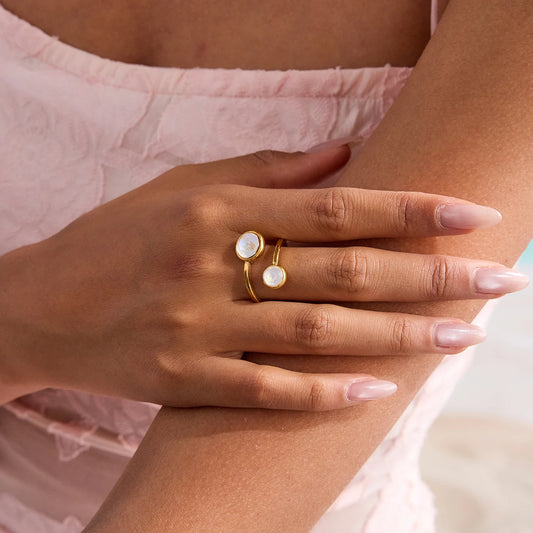 Nahaufnahme einer Hand mit gepflegten Fingernägeln, die einen offenen Wickelring aus 18K vergoldetem Edelstahl mit zwei runden, weiß schimmernden Elementen trägt.