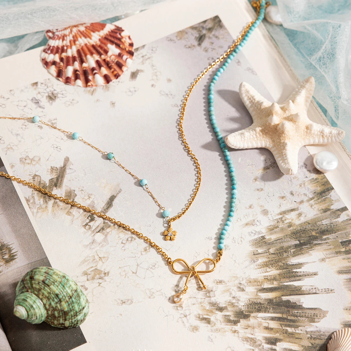 Goldene Halskette mit türkisfarbenen Perlen und Schleifen-Anhänger, drapiert auf Muscheln und Sand. Ein stilvoller, sommerlicher Schmuck-Look.