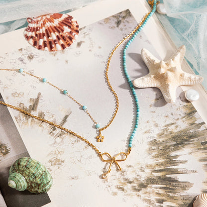 Goldene Halskette mit türkisfarbenen Perlen und Schleifen-Anhänger, drapiert auf Muscheln und Sand. Ein stilvoller, sommerlicher Schmuck-Look.