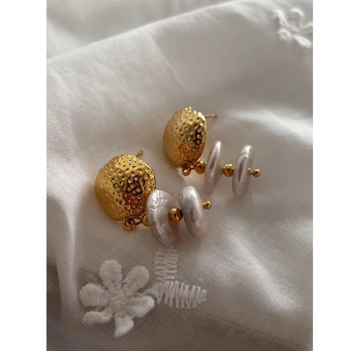 Goldene Ohrringe mit Baroque-Perlen, elegant platziert auf weißem Stoff mit einer weißen, gestickten Blumenapplikation.