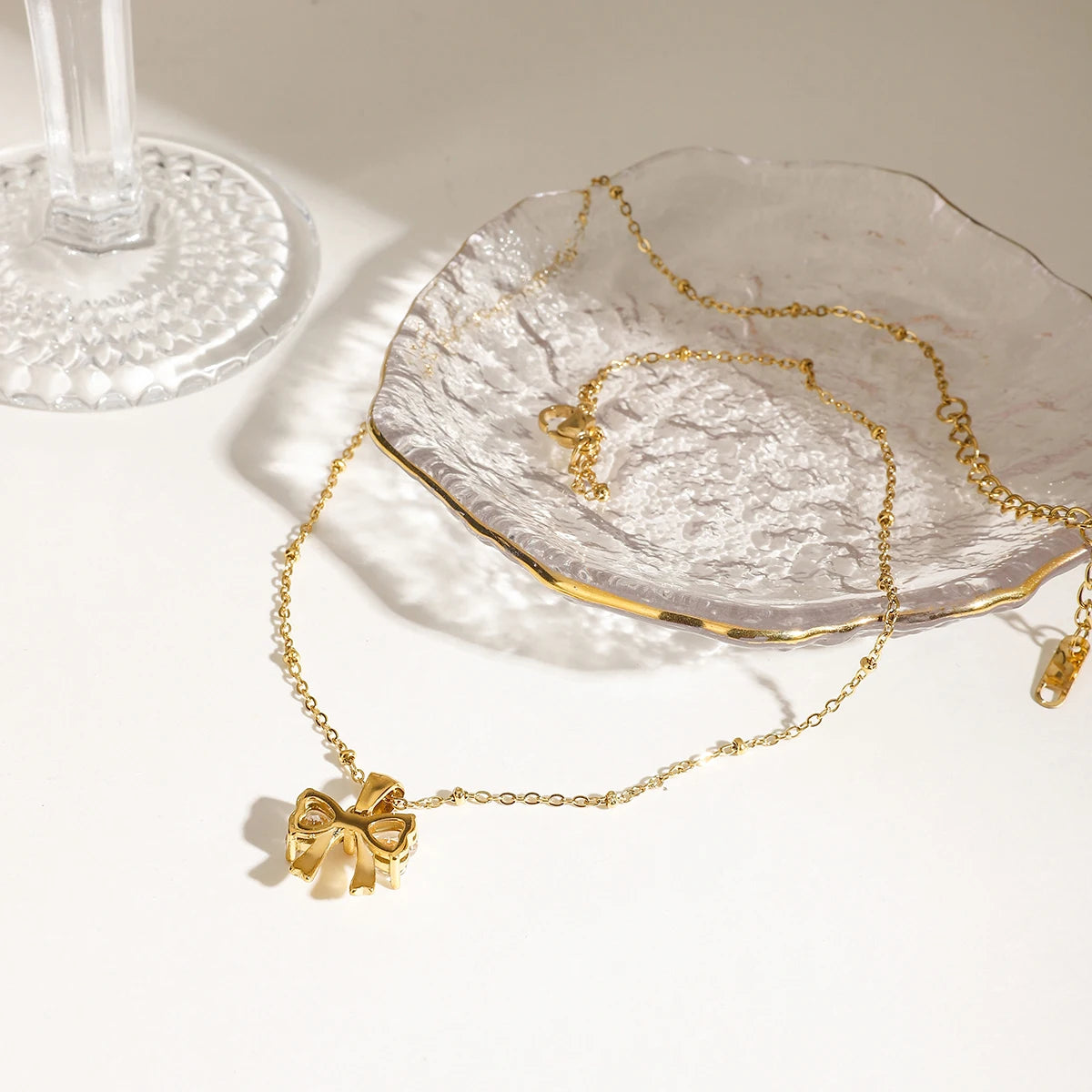 Rückseite einer goldene Edelstahlkette mit Zirkonia-Schleifenanhänger elegant platziert neben einer Glasschale