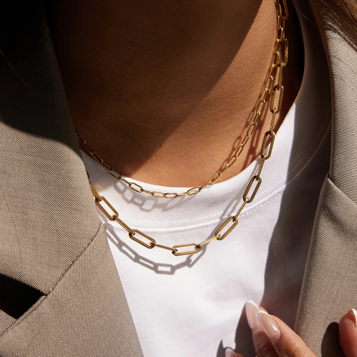 Nahaufnahme eines mehrlagigen (geschichteten) Halskettensets aus goldfarbenem Edelstahl am Hals einer Frau; die Kette besteht aus einer feineren Gliederkette und einer breiteren Gliederkette im Paperclip-Design mit 18K Gold Vergoldung, getragen über einem weißen T-Shirt und unter einem beige Blazer.