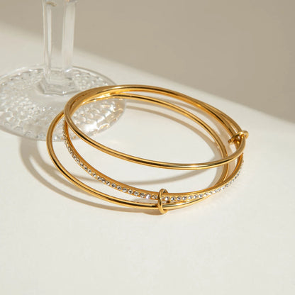 „Goldfarbener Edelstahl-Armreif mit ineinander verschlungenen Schienen und glitzerndem Steinbesatz, hochwertig 18K vergoldet.“