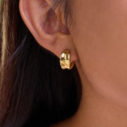Detaillierte Nahaufnahme des goldvergoldeten Edelstahl-Creolen-Ohrrings, direkt am Ohr der Trägerin, um das geschwungene, modern-organische Design und die glänzende Oberfläche zu präsentieren.