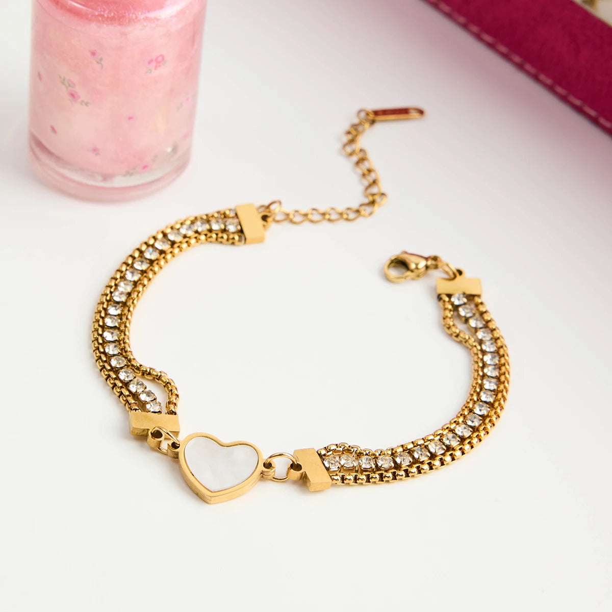 Flachbild-Produktfoto eines dreireihigen Gold-Armbands aus Edelstahl mit Strass und weißem Herz-Anhänger, arrangiert neben einer rosa Flasche.