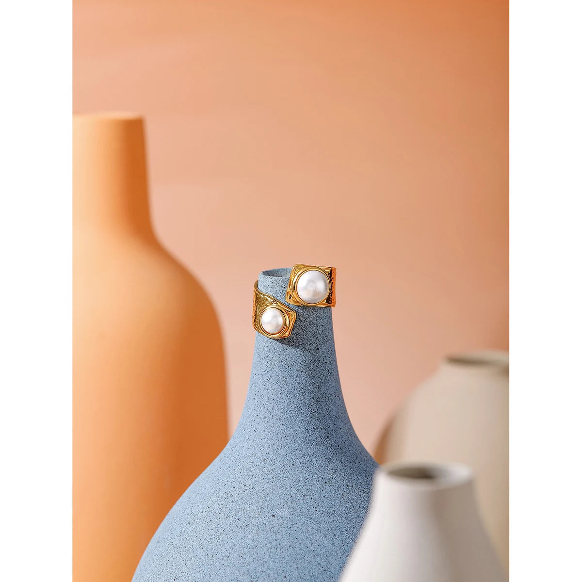 Elegantes Stillleben-Foto der vergoldeten Edelstahlringe mit Perlen, kunstvoll auf dem Flaschenhals einer blauen Steingut-Vase in einer modernen Interieur-Szenerie drapiert.